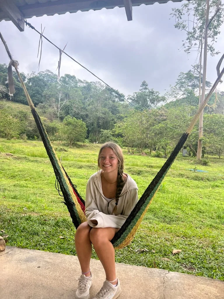 Travel agent specializing in once in a lifetime memorable trips and travel for groups. She is sitting in a colorful hammock, smiling in a lush green outdoor setting, reflecting the essence of travel and exploration.